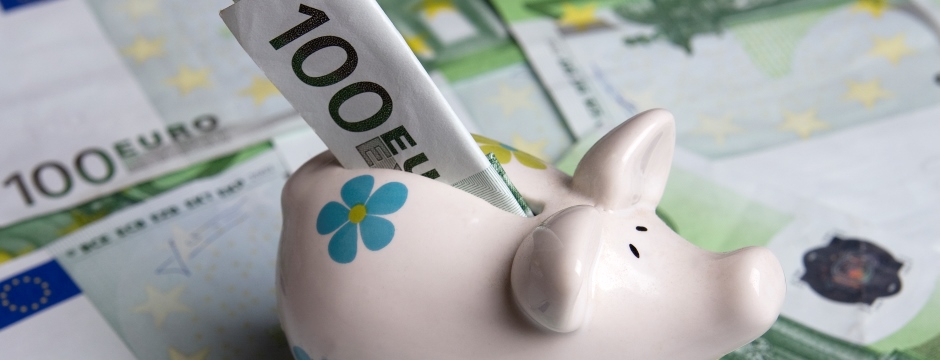 rosa Sparschwein mit blauem Blümchen und 100-Euro-Schein im Schlitz steht auf weiteren  100-Euro-Scheinen