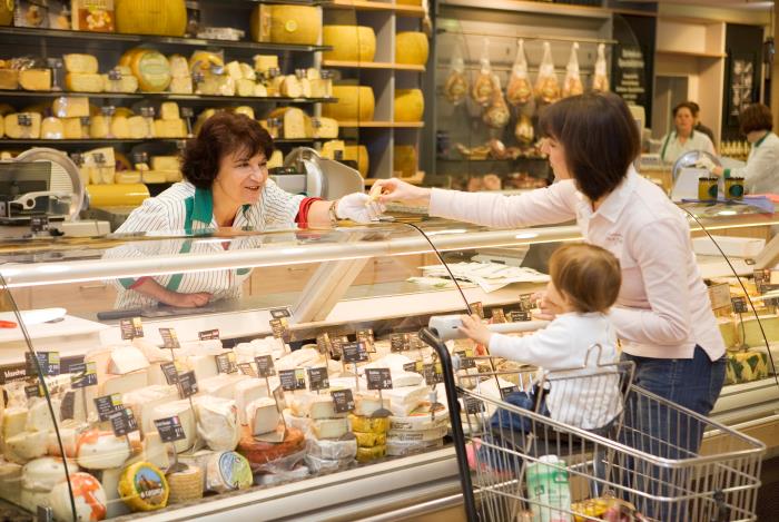 Dunkelhaarige Mutter mit Kind im Einkaufswagen wird an einer Käsetheke im Supermarkt von Verkäuferin bedient