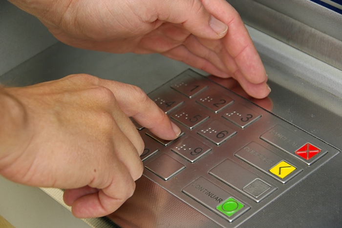 rechte Hand gibt mit Zeigefinger am Geldautomat Code in die Tastatur ein, während die linke Hand abschirmt