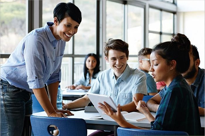 Schulklasse mit 5 Schülern und Lehrerin vor einer Wand aus Glasfenstern , alle grinsen, alle haben dunkle Haare