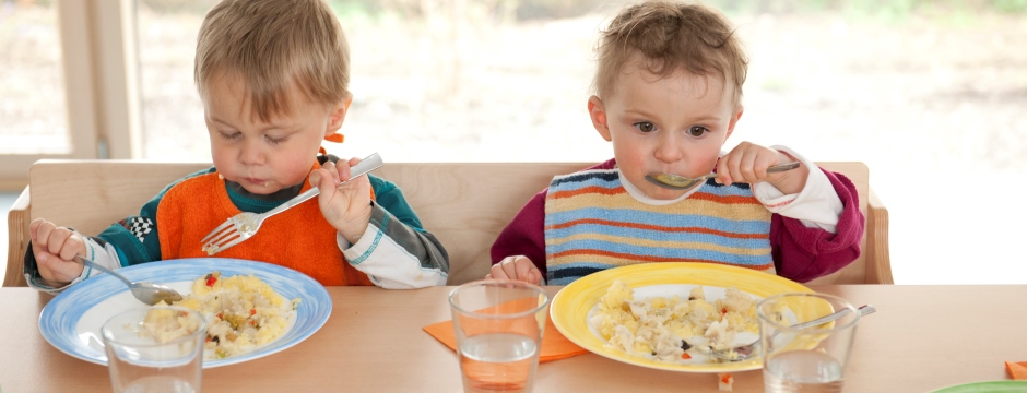 Zwei Kleinkinder essen an einem Tisch mit Besteck aus bunten Tellern, davor stehen Wassergläser und Gläser mit Rohkost gemüse