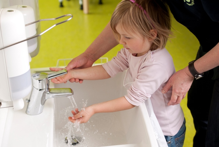 Kleines Kind mit hochgekrempelten Ärmeln steht am Waschbecken und wäscht  die Hände, beaufsichtigt von einer erwachsenen Person in schwarzer Kleidung
