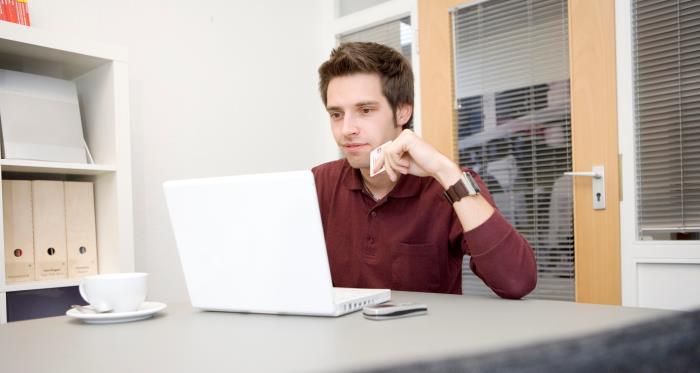 Junger Mann sitzt mit der Kreditkarte in der Hand an Bürotisch neben Kaffeetasse am Laptop und schaut hinein.