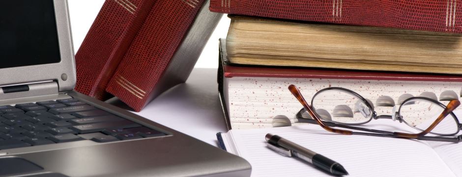 Schreibtisch-Nahaufnahme mit Brille, Kugelschreiber, Büchern und dem Teilausschnitt eines Laptops