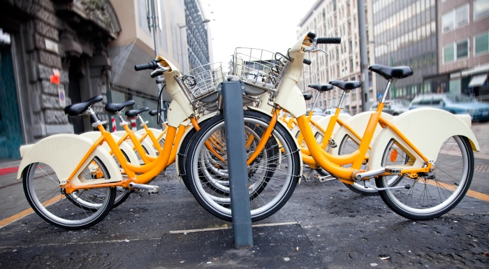 Mehrere Mietfahrrader mit gelben Rahmen stehen in einer Straße abgestellt in einer Reihe.