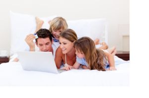 Vierköpfige Familie mit weißem Laptop in weißem Bett. Papa hat eine Kreditkarte in der Hand