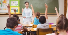 Schwarzhaarige Lehrerin in türkiserKleidung vor einer Tafel im Klassenzimmer ; vor ihr mit dem Rücken zur zum Betrachter sich meldende Kinder