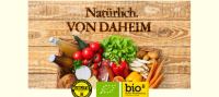Logo der Kampagne Natürlich von Daheim: Korb voll mit bunten Lebensmitteln auf Holzbretter-Hintergrund.